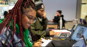 A black woman smiles as she finishes a writing assignment. She is surrounded by other working scholars.