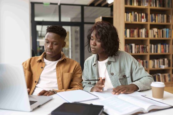 Two black students look at a laptop together, surrounded by books